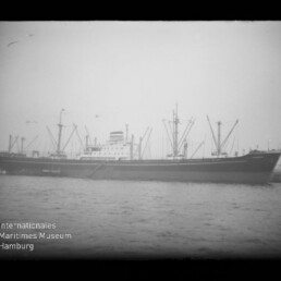 Schwarzweißaufnahme eines langen Frachtschiffs im Hafen mit mehreren Ladekränen und Aufbauten in Mittschiff, Wasser im Vordergrund und dem Logo des Internationalen Maritimen Museums Hamburg unten links.