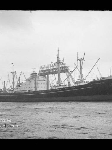 Schwarzweißaufnahme eines großen Frachtschiffs mit Ladebäumen und mehreren Masten, seitlich im Hafen auf ruhigem Wasser liegend, im Hintergrund Hafenkräne und Industrieanlagen.