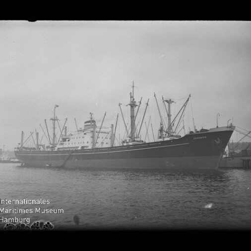 Schwarzweiß-Foto eines großen Frachtschiffs mit zahlreichen Masten und Ladekränen, vertäut am Kai eines Hafens; links unten sichtbar das Logo und der Schriftzug des Internationalen Maritimen Museums Hamburg.