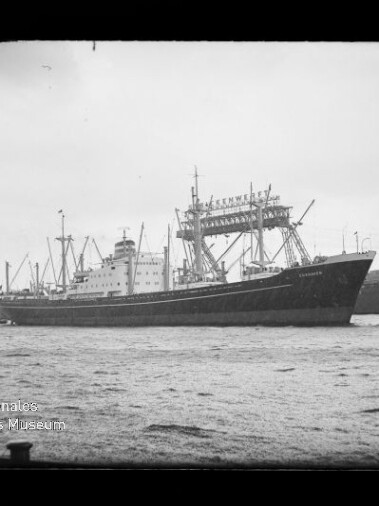 Schwarzweiß-Foto eines großen Frachtschiffs mit mehreren Ladekränen und sichtbaren Aufbauten, das in einem Industriehafen mit hohen Portalkränen im Hintergrund fährt; im Vordergrund Wasser und unten links das Logo des Internationalen Maritimen Museums Hamburg.