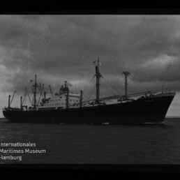 Schwarzweiß-Foto eines großen Frachtschiffs mit mehreren Ladekränen und sichtbaren Aufbauten, das bei bewölktem Himmel auf ruhigem Meer fährt; Filmrand mit Beschriftung ‚ADOX R 21‘ und Logo des Internationalen Maritimen Museums Hamburg im Bildrand.
