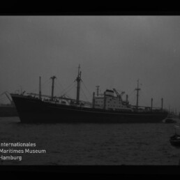 Schwarzweißaufnahme eines großen Frachtschiffs mit Ladekränen und Masten im Hafen, von kleineren Booten begleitet; unten links das Logo und der Schriftzug des Internationalen Maritimen Museums Hamburg.