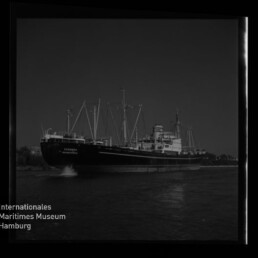 Großes Frachtschiff mit mehreren Masten und Ladekränen fährt auf ruhigem Wasser bei dunklem Himmel, mit sichtbarer Bugwelle, Filmrahmenrand und Wasserzeichen des Internationalen Maritimen Museums Hamburg unten links.