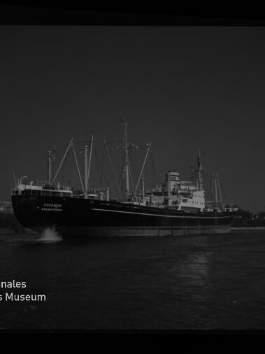 Großes Frachtschiff mit mehreren Masten und Ladekränen fährt auf ruhigem Wasser bei dunklem Himmel, mit sichtbarer Bugwelle, Filmrahmenrand und Wasserzeichen des Internationalen Maritimen Museums Hamburg unten links.