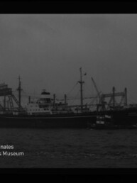 Schwarzweißaufnahme eines größeren Frachtschiffs auf dem Wasser mit einem kleinen Schlepper entlang der Seite, im Hintergrund Hafenkräne und Industrieanlagen; Museumssignet ‚Internationales Maritimes Museum Hamburg‘ unten links.