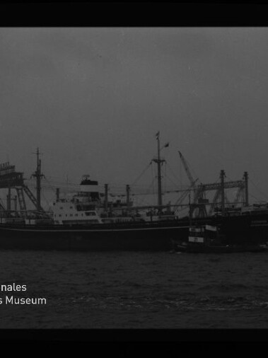 Schwarzweißaufnahme eines größeren Frachtschiffs auf dem Wasser mit einem kleinen Schlepper entlang der Seite, im Hintergrund Hafenkräne und Industrieanlagen; Museumssignet ‚Internationales Maritimes Museum Hamburg‘ unten links.