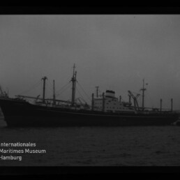 Schwarzweißaufnahme eines langen Frachtschiffs mit mehreren Ladekränen und Masten, im Hafen vor dunklem Wasser liegend, mit sichtbarem Filmrand und Museumssignet in der linken unteren Ecke.