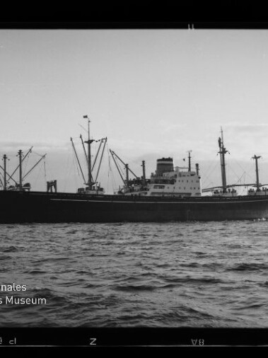 Schwarzweißaufnahme eines Frachtschiffs mit mehreren Ladekränen und mittig gelegenem Aufbau auf ruhiger See vor flachem Horizont; Filmrand und Wasserzeichen des Internationalen Maritimen Museums Hamburg in der unteren linken Ecke sichtbar.