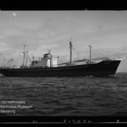 Schwarzweiß-Foto eines Frachtschiffs in Seitenansicht auf ruhigem Meer mit mehreren Ladekränen und mittigem Aufbauten; unten links das Logo des Internationalen Maritimen Museums Hamburg.
