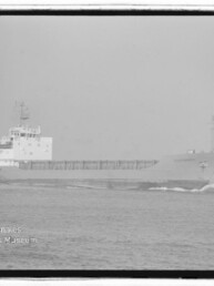 Schwarzweißfoto eines Frachtschiffs, das von links nach rechts über eine ruhige See fährt; sichtbar sind Bugwelle, die lange offene Ladungsbucht und die Aufbauten am Heck sowie ein Wasserzeichen des Internationalen Maritimen Museums Hamburg unten links.