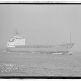 Schwarzweißfoto eines Frachtschiffs, das von links nach rechts über eine ruhige See fährt; sichtbar sind Bugwelle, die lange offene Ladungsbucht und die Aufbauten am Heck sowie ein Wasserzeichen des Internationalen Maritimen Museums Hamburg unten links.