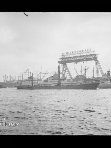 Frachtschiff im Hafen vor einer Werft mit großem Portalkran und dem Schriftzug Stülckenwerft, im Hintergrund weitere Kräne und Schiffe und im Vordergrund die Wasseroberfläche.