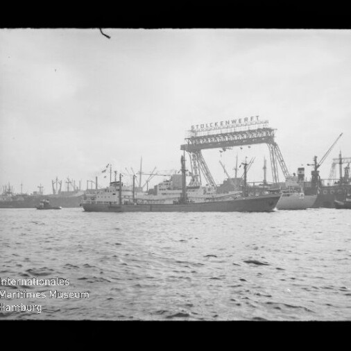 Frachtschiff im Hafen vor einer Werft mit großem Portalkran und dem Schriftzug Stülckenwerft, im Hintergrund weitere Kräne und Schiffe und im Vordergrund die Wasseroberfläche.
