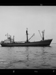 Schwarzweißfoto eines Frachtschiffs mit Ladebäumen und Decksaufbauten, seitlich im ruhigen Meer vor flachem, niedrigem Horizont, umgeben von sichtbarem Filmrand und dem Logo des Internationalen Maritimen Museums Hamburg.