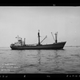 Schwarzweißfoto eines Frachtschiffs mit Ladebäumen und Decksaufbauten, seitlich im ruhigen Meer vor flachem, niedrigem Horizont, umgeben von sichtbarem Filmrand und dem Logo des Internationalen Maritimen Museums Hamburg.