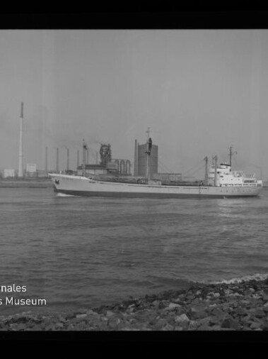 Frachtschiff fährt auf ruhigem Wasser vor einem Industriehafen mit Silotürmen, Schornstein und Krananlagen; im Vordergrund ein steiniger Uferstreifen und links unten das Logo des Internationalen Maritimen Museums Hamburg.