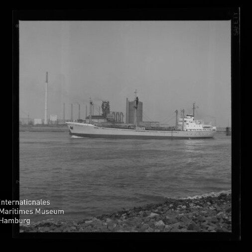 Frachtschiff fährt auf ruhigem Wasser vor einem Industriehafen mit Silotürmen, Schornstein und Krananlagen; im Vordergrund ein steiniger Uferstreifen und links unten das Logo des Internationalen Maritimen Museums Hamburg.