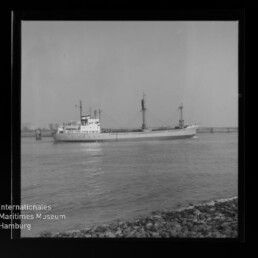 Schwarz-weiß-Foto eines weiß gestrichenen Frachtschiffs mit zwei Ladebaum-Masten, das auf ruhigem Wasser vor einem flachen Ufer mit Steinböschung fährt; links unten das Logo des Internationalen Maritimen Museums Hamburg.