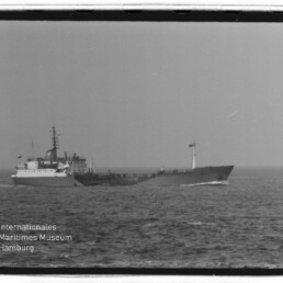 Schwarz-weiß-Foto eines langen, niedrig bauenden Frachtschiffs mit Aufbauten mittschiffs, Masten und Flagge am Bug, das auf ruhiger See fährt; unten links das Logo des Internationalen Maritimen Museums Hamburg.