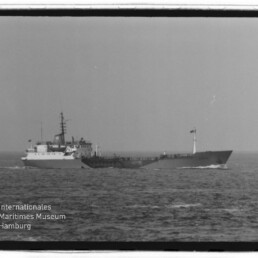 Schwarzweißaufnahme eines Frachtschiffs auf ruhiger See mit niedrigem Deckaufbau, hohem Funkmast und Flagge; unten links sichtbar das Wasserzeichen 'Internationales Maritimes Museum Hamburg'.