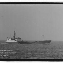 Schwarzweißaufnahme eines mittleren Frachtschiffs auf offener See mit auffälliger Brücke achtern, sichtbarer Flagge am Bug und leichtem Wellengang; Foto zeigt auch ein Wasserzeichen „Internationales Maritimes Museum Hamburg“ unten links.