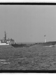 Schwarzweißaufnahme eines seitlich fahrenden Frachtschiffs/Tankers auf ruhiger See mit deutlich sichtbarem Brückenaufbau, Mast und Flagge; unten links Logo des Internationalen Maritimen Museums Hamburg.