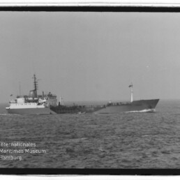 Schwarzweißaufnahme eines seitlich fahrenden Frachtschiffs/Tankers auf ruhiger See mit deutlich sichtbarem Brückenaufbau, Mast und Flagge; unten links Logo des Internationalen Maritimen Museums Hamburg.