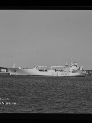 Schwarzweißaufnahme eines großen Frachtschiffs mit der Aufschrift „OOCL CARRIERS“, das auf ruhigem Wasser vor einer Küstenlinie mit Gebäuden und Pier fährt; links unten Museumssignet und Schriftzug des Internationalen Maritimen Museums Hamburg sowie schwarzer Filmrand mit Markierung.