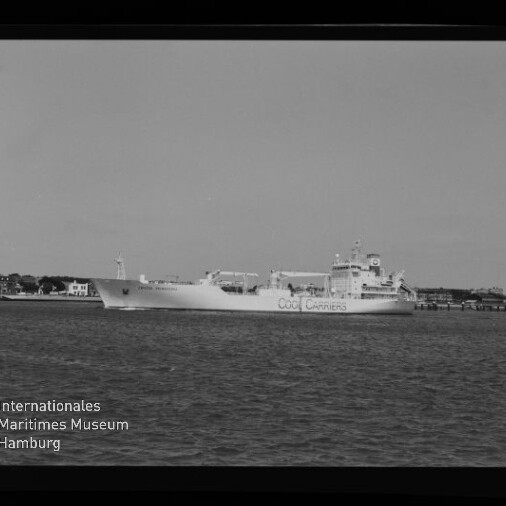 Schwarzweißaufnahme eines großen Frachtschiffs mit der Aufschrift „OOCL CARRIERS“, das auf ruhigem Wasser vor einer Küstenlinie mit Gebäuden und Pier fährt; links unten Museumssignet und Schriftzug des Internationalen Maritimen Museums Hamburg sowie schwarzer Filmrand mit Markierung.