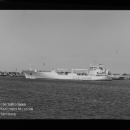 Schwarzweißaufnahme eines großen Frachtschiffs mit der Aufschrift COOL CARRIERS im Hafen, im Vordergrund Wasser, am Bildrand Filmmarkierung AGFA APX 100 und Museumslogo unten links.