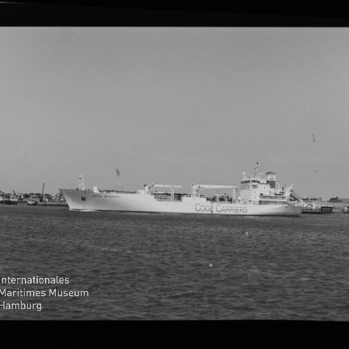 Schwarzweißaufnahme eines großen Frachtschiffs mit der Aufschrift COOL CARRIERS im Hafen, im Vordergrund Wasser, am Bildrand Filmmarkierung AGFA APX 100 und Museumslogo unten links.