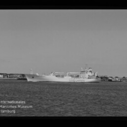 Schwarz-weiß-Foto eines großen Frachtschiffs mit dem Schriftzug „Cool Carriers“, das in ruhigem Wasser vor einer niedrigen Küstenlinie mit Gebäuden fährt; Filmrand und Wasserzeichen des Internationalen Maritimen Museums Hamburg sind sichtbar.
