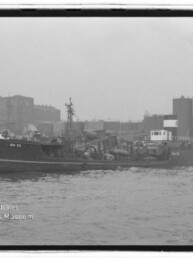 Schwarzweißfoto eines Frachtschiffs mit der Kennung DW 32, das an einer Kaimauer in einem Industriehafen vor Lagergebäuden, großen zylindrischen Tanks und rauchenden Schornsteinen liegt.