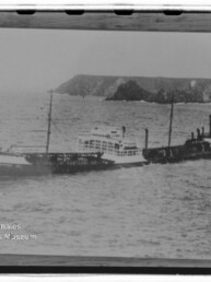 Schwarzweißaufnahme eines langen Dampfschiffs mit Masten und Schornstein, das in bewegter See nahe einer felsigen Küste fährt; unten links sichtbar das Logo und der Schriftzug des Internationalen Maritimen Museums Hamburg.