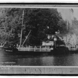 Schwarzweiß-Foto eines Dampffrachtschiffs mit zwei Masten und einem Schornstein, längsseits am Kai vor einem bewaldeten Hang und einer steilen Felswand.