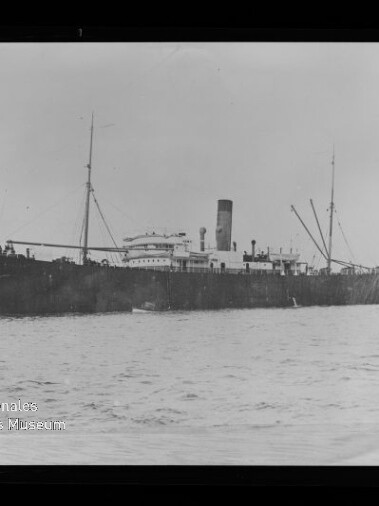 Schwarz-weißes historisches Foto einer langen Dampffrachtschiffs-Seitenansicht mit mittigem Schornstein, zwei Masten und Ladegeschirr, ruhig auf dem Meer vor hellem Himmel; links unten sichtbar das Wasserzeichen des Internationalen Maritimen Museums Hamburg und der Rand des Negativrahmens.
