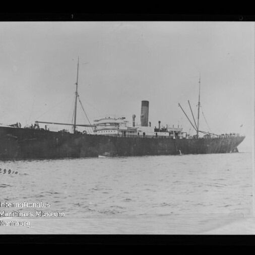 Schwarz-weißes historisches Foto einer langen Dampffrachtschiffs-Seitenansicht mit mittigem Schornstein, zwei Masten und Ladegeschirr, ruhig auf dem Meer vor hellem Himmel; links unten sichtbar das Wasserzeichen des Internationalen Maritimen Museums Hamburg und der Rand des Negativrahmens.