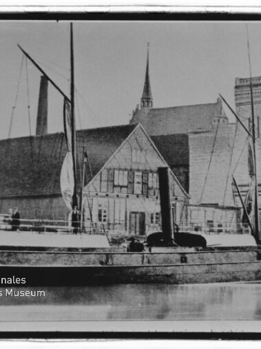 Dampfgetriebenes Schiff mit Schornstein und Masten liegt am Kai vor einem Fachwerkhaus und einem großen Backsteingebäude mit Turm; Spiegelung im ruhigen Wasser und Museumswasserzeichen unten links.
