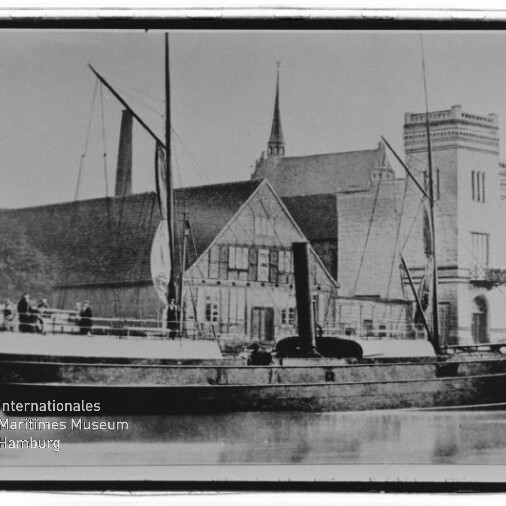 Dampfgetriebenes Schiff mit Schornstein und Masten liegt am Kai vor einem Fachwerkhaus und einem großen Backsteingebäude mit Turm; Spiegelung im ruhigen Wasser und Museumswasserzeichen unten links.