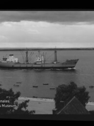 Schwarzweiß-Fotografie eines Frachtschiffs, das nahe an der Küste vorbeifährt; im Vordergrund Strand, Bäume und Hausdächer, mehrere kleine Boote auf dem Wasser, bewölkter Himmel sowie sichtbarer Filmrand und Museumswasserzeichen.