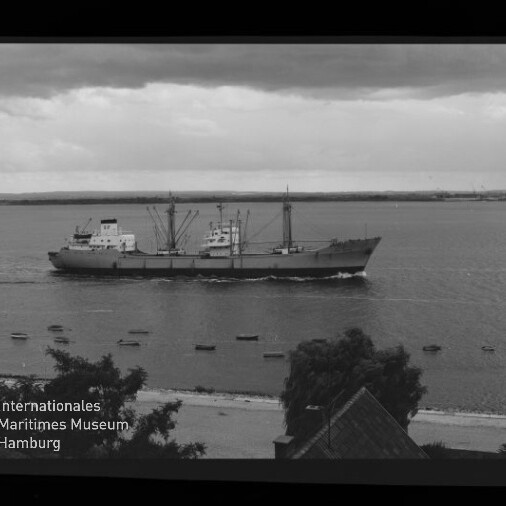 Schwarzweiß-Fotografie eines Frachtschiffs, das nahe an der Küste vorbeifährt; im Vordergrund Strand, Bäume und Hausdächer, mehrere kleine Boote auf dem Wasser, bewölkter Himmel sowie sichtbarer Filmrand und Museumswasserzeichen.