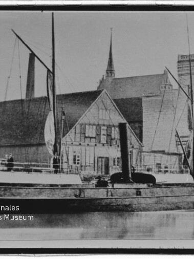 Schwarzweißaufnahme eines historischen Dampfschiffs mit Schornstein, Masten und aufgeholten Segeln, das am Kai vor einem Fachwerkgebäude und einem großen Backsteinhaus liegt, mit Spiegelung im ruhigen Wasser und einigen Personen auf dem Deck.