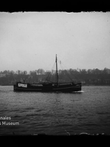 Schwarzweiß-Foto eines einmastigen Frachtschiffs, das ruhig auf dem Wasser vor einem baumbestandenen, nebligen Ufer mit Gebäuden fährt; unten links das Logo des Internationalen Maritimen Museums Hamburg.