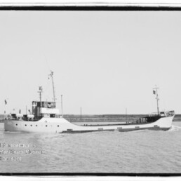 Schwarzweißaufnahme eines kleinen Küstenversorgers mit niedrigem Freibord, achterlichem Steuerhaus, Mast und langem flachen Vorschiff, der ruhig auf dem Wasser vor einer flachen Küstenlinie fährt; Museumslogo unten links sichtbar.