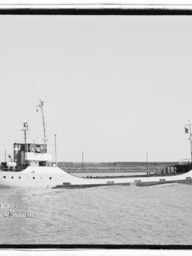 Schwarzweißaufnahme eines kleinen Küstenversorgers mit niedrigem Freibord, achterlichem Steuerhaus, Mast und langem flachen Vorschiff, der ruhig auf dem Wasser vor einer flachen Küstenlinie fährt; Museumslogo unten links sichtbar.