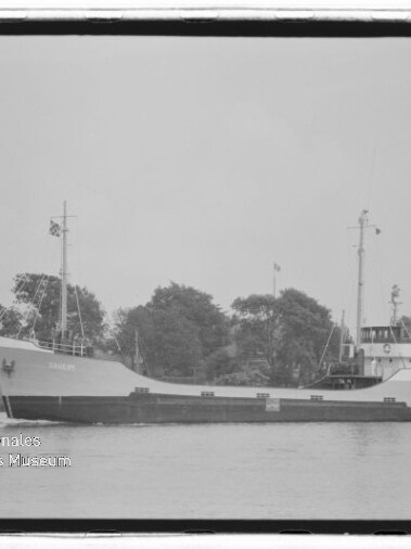 Schwarzweißaufnahme eines Frachtschiffs in Seitenansicht mit niedrigem Laderaum, zwei Masten und achterlichem Steuerhaus, vor einem baumbestandenen Ufer auf ruhigem Wasser.