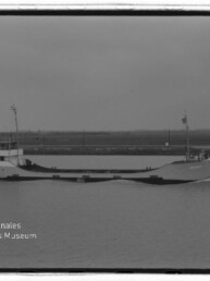 Schwarzweiß-Foto eines flachen Frachtschiffs, das in ruhigem Gewässer an einem flachen Ufer vorbeifährt; links im Vordergrund ein kleines Segelboot und in der unteren linken Ecke ein Wasserzeichen des Internationalen Maritimen Museums Hamburg.