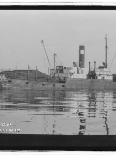 Schwarzweiß-Foto eines alten Frachtschiffs mit markantem Schornstein, mehreren Masten und Ladekränen, ruhig im Wasser liegend mit klarer Spiegelung und dem Wasserzeichen ‚Internationales Maritimes Museum Hamburg‘ links unten.