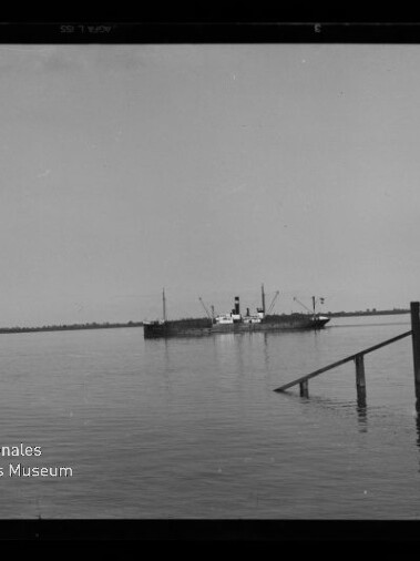 Ruhige Wasserfläche mit einem Dampfschiff in Bildmitte, einem kleinen Segelboot links und einem Pier mit einigen Personen rechts, flacher Horizont im Hintergrund.