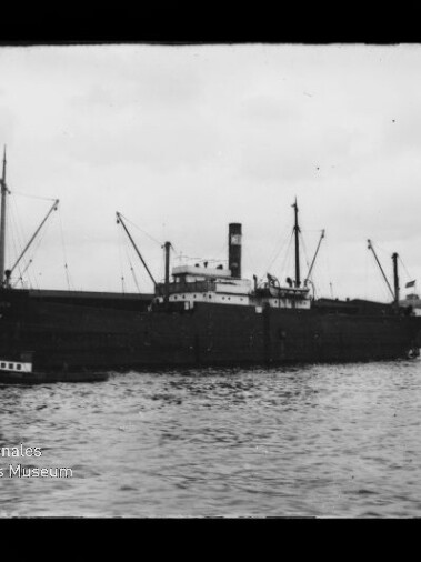Schwarzweißaufnahme eines großen Frachtschiffs mit Schornstein und Ladebaum-Masten, an einem Kai liegend, kleines Boot am Bug, Gasholder im Hintergrund und leicht bewegte Wasseroberfläche im Vordergrund.